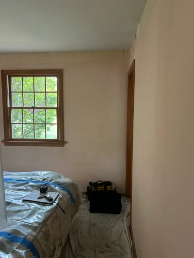 Bedroom with peach walls and wood-trimmed window, furniture covered in plastic before painting work begins.