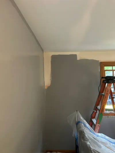 A wall midway through repainting, showing a sharp line between old off-white and new gray paint near a ladder.
