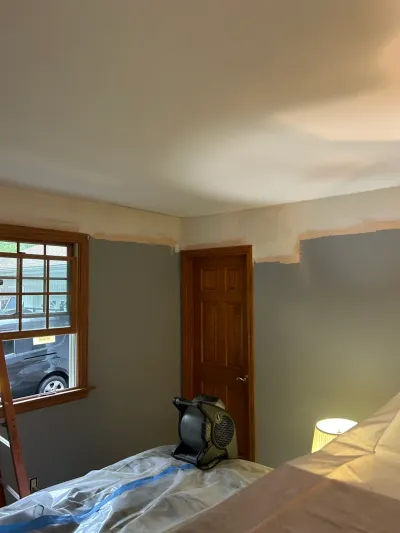 A bedroom mid-repaint with gray walls in progress, drop cloths, and a fan drying the paint.