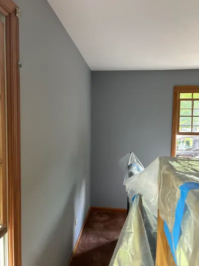 A room mid-repaint with fresh gray-blue walls, wood trim, and furniture covered in plastic sheeting.
