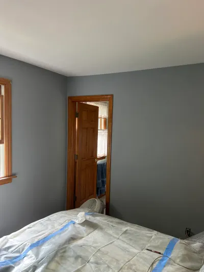 A bedroom mid-painting with gray walls, a wood door ajar, and furniture covered in plastic sheeting.