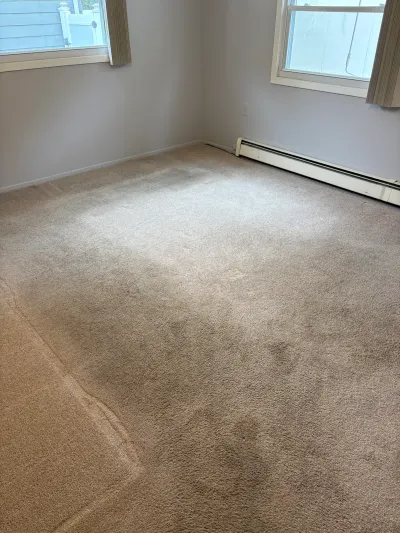 Empty bedroom with worn, stained beige carpet and two corner windows before renovation.