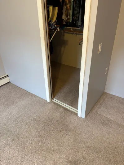 Open sliding closet door revealing hanging clothes in a carpeted bedroom.