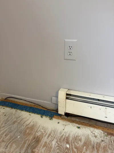 Room mid-renovation showing exposed plywood subfloor, old carpet remnant, and a baseboard heater with hanging wires.