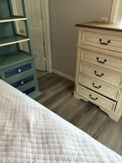 Bedroom corner with a sage green nightstand and a distressed cream dresser on wood-look flooring.