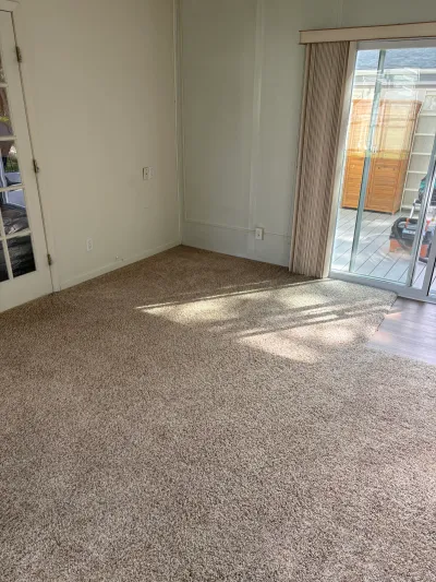 Empty carpeted living room with a sliding glass door leading to a deck and a french door on the side wall.