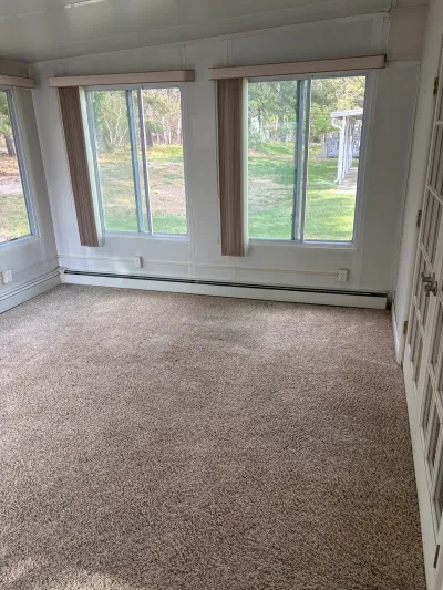 Empty sunroom with worn carpet, baseboard heater, and large windows overlooking a grassy yard.