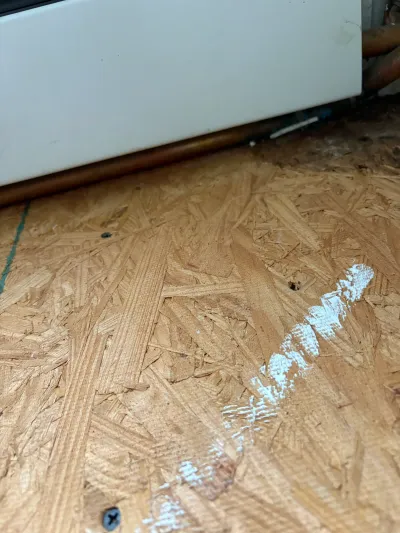 Close-up of exposed OSB subfloor with a copper pipe and white paint smear during a remodel.