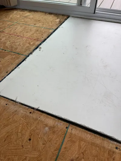 Close-up of a subfloor transitioning from OSB plywood to white tile backer board near a sliding door.