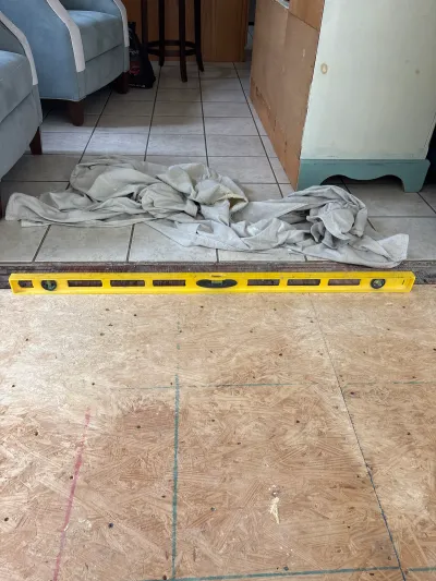 A level tool lies across the seam between old tile flooring and new plywood subfloor during a renovation.