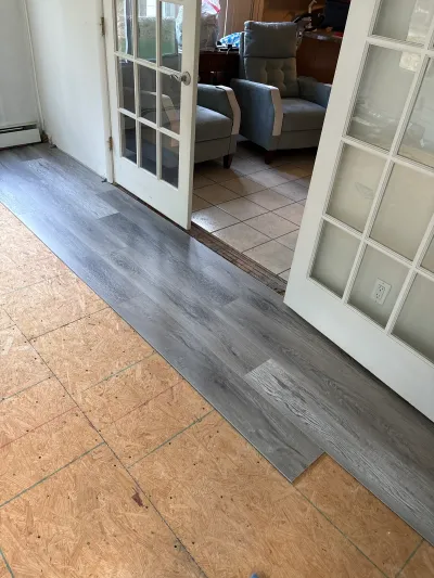 New grey vinyl plank flooring being installed over subfloor, with French doors opening to a tiled room beyond.