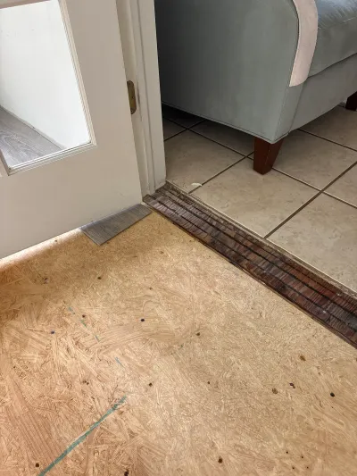 Bare plywood subfloor at an entryway mid-renovation, with a tile room and sofa visible beyond the door.