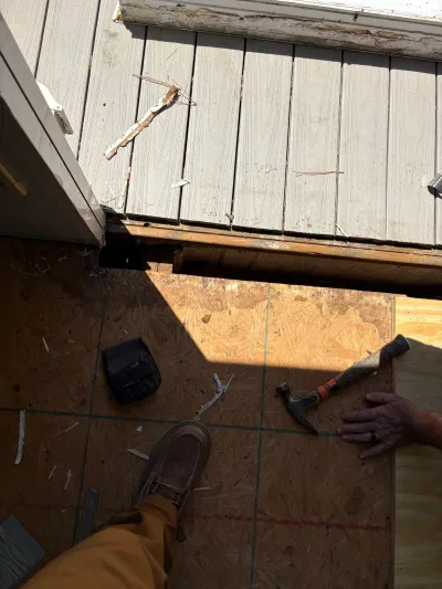 Overhead view of a construction worker's hand and foot on a plywood subfloor near damaged exterior siding.