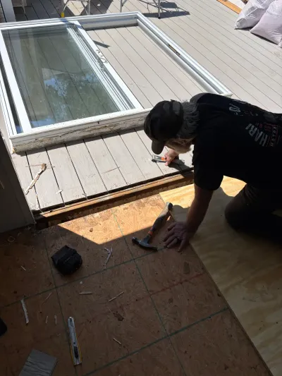 Worker kneeling on new plywood subfloor during a deck renovation, with an old window frame propped nearby.
