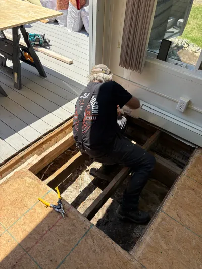 Contractor repairs exposed floor joists in a sunroom with subfloor panels removed.