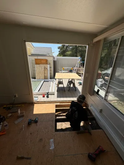 A worker kneels in a floor cutout during construction of a sunroom that opens onto an unfinished deck.