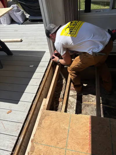 Worker repairing a deck by drilling into exposed framing next to a cutout section of decking.