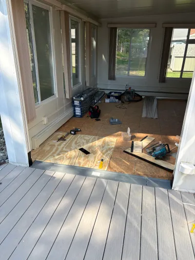 A sunroom mid-renovation with exposed subfloor, tools, and stacked flooring boxes, seen from an adjoining deck.