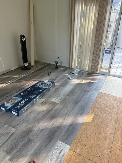Living room floor mid-installation with grey vinyl plank flooring meeting exposed plywood subfloor.