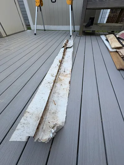 A rusted, damaged length of gutter trim removed and laid on a composite deck near sawhorses and scrap wood.
