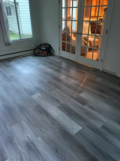 A room with new grey plank flooring being installed, a shop vacuum in the corner, and French doors to a sunroom.