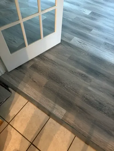 Close-up of new grey wood-look vinyl plank flooring meeting tile at a white paned glass door threshold.
