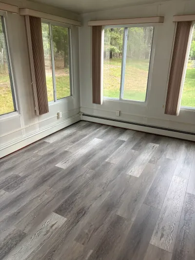Empty sunroom with new grey wood-look vinyl plank flooring and large windows overlooking a grassy yard.