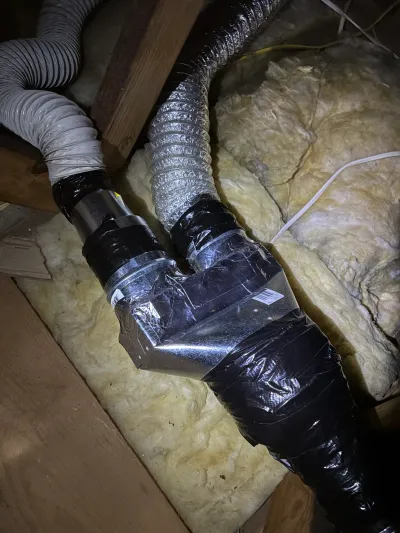 Two flexible ducts join at a metal Y-fitting in an attic, surrounded by insulation and wood framing.