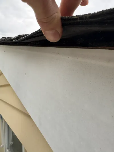 Close-up of a hand lifting a shingle edge to inspect roofline condition above fascia and siding.