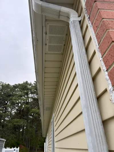 Close-up of a house corner where cream vinyl siding meets brick, with a gutter downspout and soffit vent.