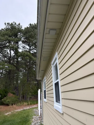 Close-up angle of a house exterior showing tan vinyl siding, white-trimmed windows, and roof soffit with gutter.