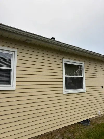 Side view of a house exterior with tan vinyl siding, white-trimmed windows, and a gutter along the roofline.