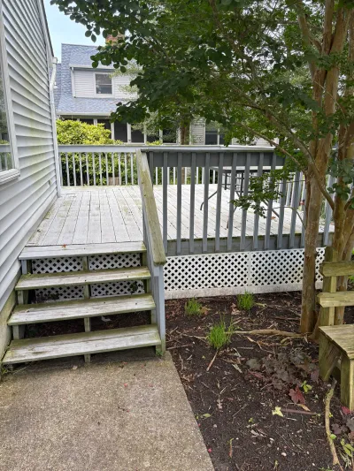 Weathered wood deck and stairs leading to a concrete walkway beside a house, with a garden bed nearby.