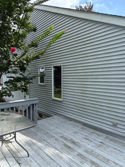 Side exterior of a house with gray vinyl siding, seen from a weathered wood deck with a patio table.