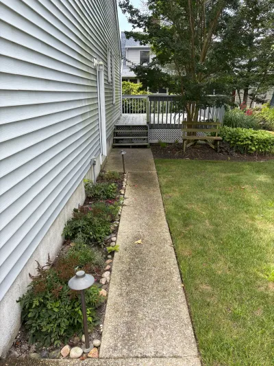 Concrete side walkway between a house and lawn leading to a wood deck with landscaped garden beds.