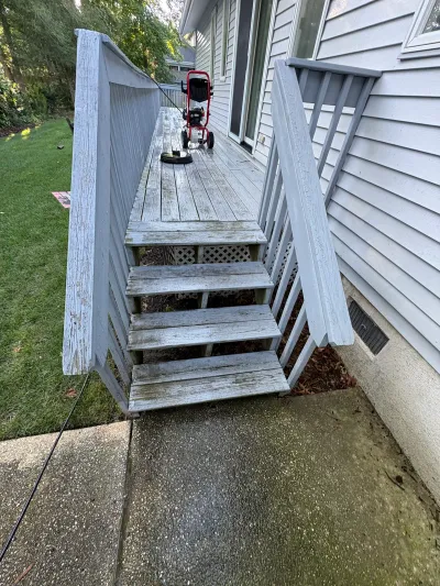 A weathered wood deck and stairs mid pressure-washing, with peeling paint and a washer on the deck.oz