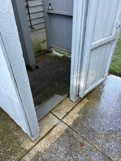 Weathered outdoor shed doors open to reveal wood flooring and a stained stucco wall with an exterior spigot.