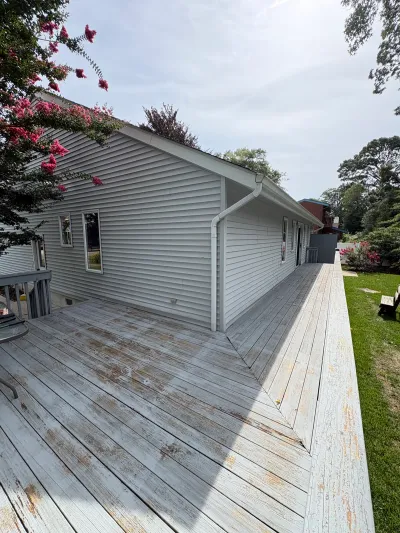Weathered side deck with peeling paint runs along a gray vinyl-sided house under an overcast sky.