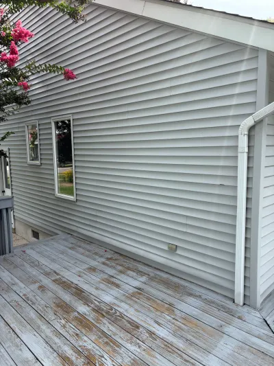 Exterior wall with grey vinyl siding above a weathered wood deck, with a flowering tree overhead.