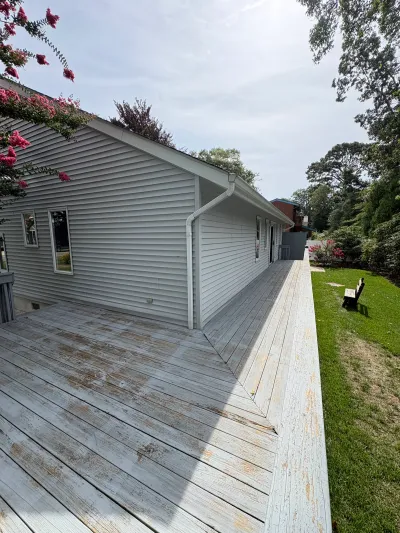 Weathered wood deck runs alongside a grey-sided house with peeling railing paint and a flowering tree overhead.