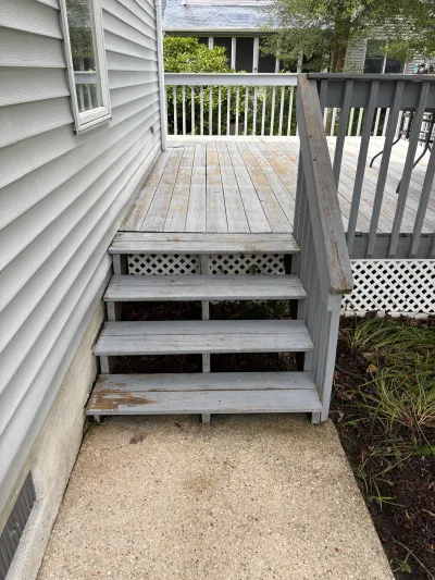 Weathered wooden deck stairs with peeling finish leading down to a concrete walkway beside the house.