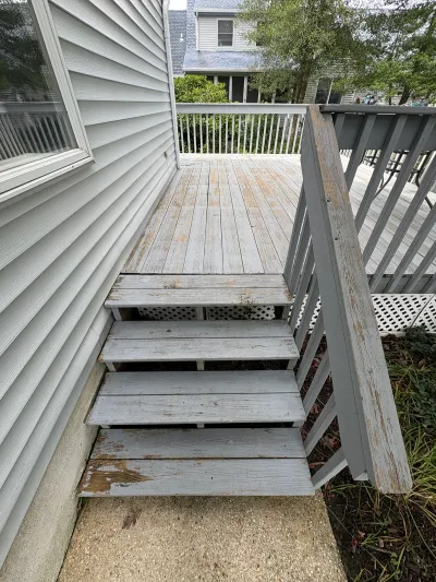Weathered wooden deck stairs with peeling paint leading down to a concrete landing beside the house.