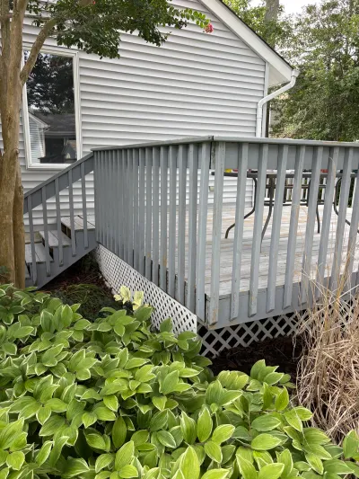 Weathered grey deck with peeling paint and a staircase, seen past hostas and lattice skirting.