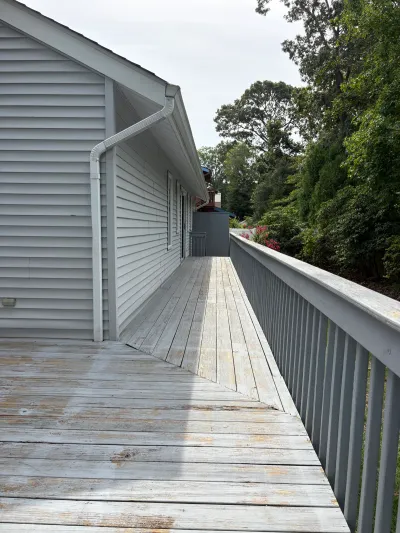 A weathered wood deck runs along the side of a grey-sided house, bordered by trees and shrubs.