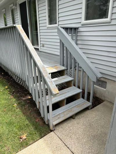 Weathered wooden deck stairs with peeling paint leading down to a concrete patio beside a house.