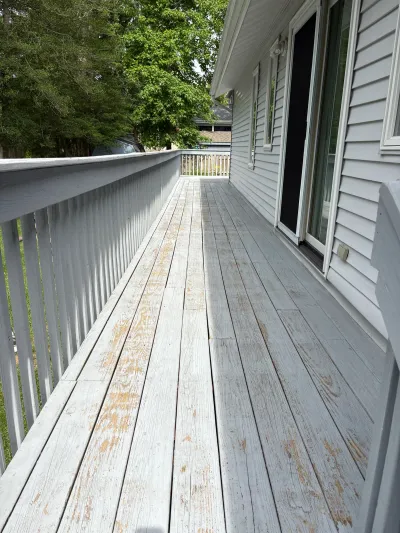 A weathered deck with peeling grey paint runs alongside a house with vinyl siding and a sliding door.
