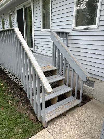 Weathered wooden deck stairs with peeling grey paint leading to a concrete walkway beside a house.