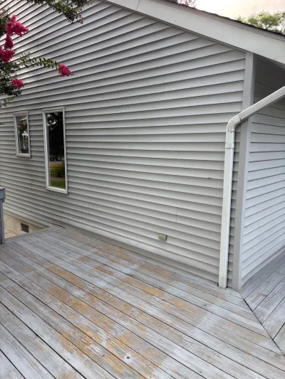 Weathered wood deck beneath grey vinyl siding with two narrow windows and a downspout.