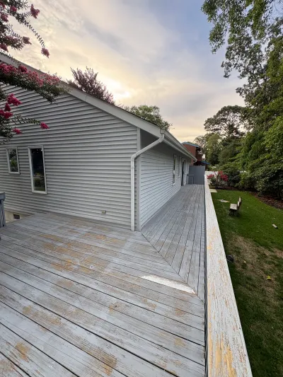 Weathered wood deck with peeling paint along the side of a grey vinyl-sided house before renovation.