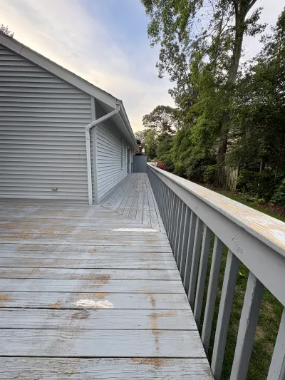 Weathered wood deck with peeling paint running alongside a house with grey vinyl siding.